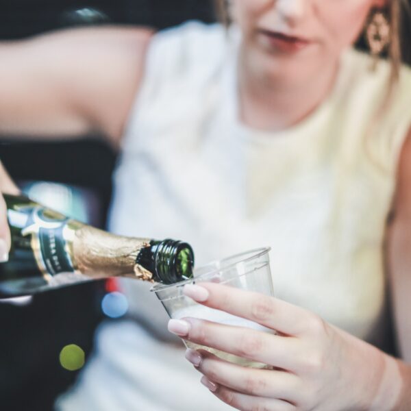 Eine Frau in weißer Kleidung gießt Champagner aus einer Flasche in einen Plastikbecher ein, wobei sie sowohl den Becher als auch die Flasche hält, wobei der Hintergrund unscharf ist.