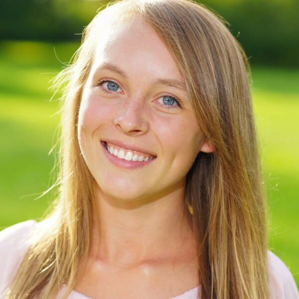 Eine junge Frau mit glattem blondem Haar und blauen Augen lächelt in die Kamera, während sie an einem sonnigen Tag im Freien steht, mit grünem Gras und verschwommenen Bäumen im Hintergrund.