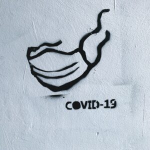 Schwarzes Schablonengraffiti einer Gesichtsmaske und des Textes COVID-19 auf einer rauen weißen Wand, das die Coronavirus-Pandemie und das Maskentragen symbolisiert.
