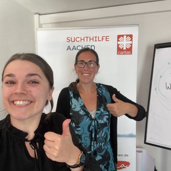 Zwei lächelnde Frauen zeigen den Daumen nach oben vor einem Transparent mit der Aufschrift "Suchthilfe Aachen" und einem Flipchart mit dem handgeschriebenen Text "Herzlich Willkommen" und einem roten Herz. Sie scheinen sich in einem Büro oder Besprechungsraum zu befinden.
