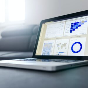 Ein Laptop auf einer Couch, auf dem Datenanalyse-Dashboards mit Grafiken, Diagrammen, einer Weltkarte und einem Kreisdiagramm angezeigt werden, während im Hintergrund das Sonnenlicht durch ein Fenster fällt.