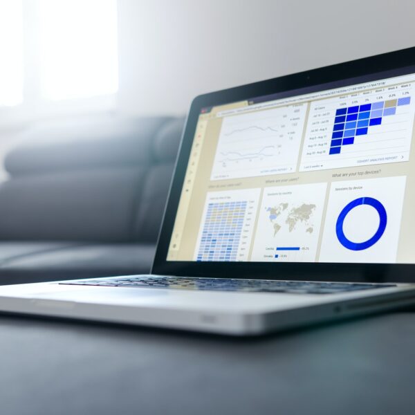 Ein Laptop auf einer Couch, auf dem Datenanalyse-Dashboards mit Grafiken, Diagrammen, einer Weltkarte und einem Kreisdiagramm angezeigt werden, während im Hintergrund das Sonnenlicht durch ein Fenster fällt.