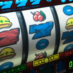 Eine Nahaufnahme eines Spielautomaten-Displays zeigt Symbole eines gelben Helms, rote und blaue Gegenstände, Kirschen und das Wort JAC auf den Walzen, mit etwas japanischem Text und bunten Grafiken rund um den Bildschirm.