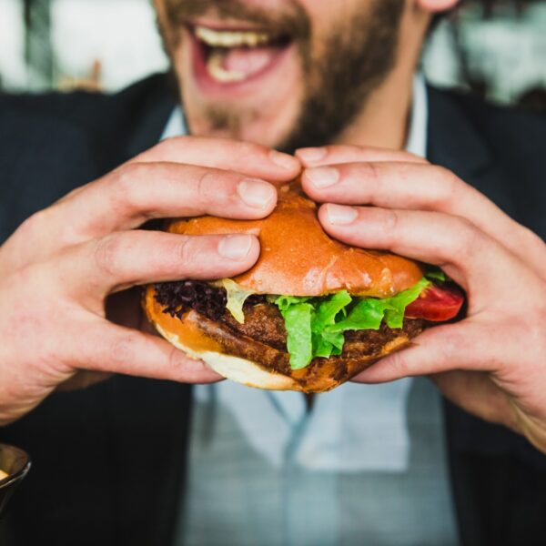 Eine lächelnde Person, die einen großen Hamburger mit Salat, Tomate und Käse in der Hand hält und sich darauf vorbereitet, einen Bissen zu nehmen. Im Vordergrund ist eine Schüssel mit Pommes frites zu sehen.