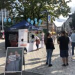 Eine Straßenszene mit Menschen, die sich um ein blaues Zelt mit Bannern und Luftballons versammelt haben. Im Vordergrund ist ein großes Plakat zu sehen, im Hintergrund Gebäude, Bäume und Fußgänger an einem sonnigen Tag.