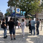 Menschen versammeln sich um einen Informationsstand mit blauen Luftballons auf einem Stadtplatz. Einige unterhalten sich, während andere auf die Anzeige schauen. Im Hintergrund sind Gebäude, Bäume und ein Straßenschild zu sehen.