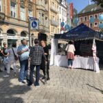 Eine Straßenszene zeigt Menschen, die sich in der Nähe eines blauen Zeltes mit Luftballons und Informationsschildern versammelt haben. Einige Leute unterhalten sich, während andere das Zelt besuchen. Historische Gebäude und Geschäfte säumen den Hintergrund an einem sonnigen Tag.