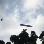 Ein kleines Flugzeug fliegt am wolkenverhangenen Himmel über den Baumwipfeln, im Schlepptau ein Transparent mit der Aufschrift fragEltern.de.