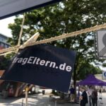 Eine dunkelblaue Karte mit dem Text fragEltern.de ist mit einer Wäscheklammer an einem Seil befestigt. Im Hintergrund laufen Menschen auf einem Stadtplatz, auf dem Bäume, Zelte und ein Straßenschild zu sehen sind.
