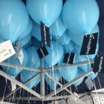 Blaue Luftballons unter einem Zeltdach, die mit Schnüren zusammengebunden und an weißen oder dunklen Anhängern mit der Aufschrift "frageEltern.de" befestigt sind. Auf einigen Schildern sind handschriftliche Botschaften auf Deutsch zu lesen.