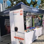 Ein Straßenstand mit blauen Luftballons und Bannern zur Drogenprävention. Unter einem blauen Zelt mit dem Schriftzug fragEltern.de liegen Informationsplakate und Broschüren aus, darunter eine mit der Aufschrift SuchtHilfe Aachen.