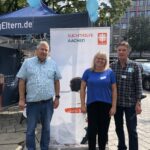 Drei Personen stehen bei einer Veranstaltung im Freien vor einem Banner der Suchthilfe Aachen. Blaue Luftballons schweben über ihnen, im Hintergrund sind ein blaues Zelt und Bäume zu sehen.