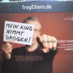 Eine Person hält ein Schild mit der Aufschrift MEIN KIND NIMMT DROGEN! vor einem Transparent mit den Aufschriften fragEltern.de und Du denkst: Selbst schuld? Drogen kommen in die besten Familien vor.