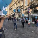 Blaue Ballons schweben in den teilweise bewölkten Himmel, umrahmt von den Spitzen zweier Gebäude - ein modernes und ein verziertes mit Bögen und Backsteinverzierungen.