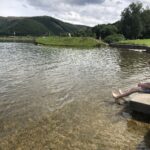 Eine Person sitzt mit nackten Füßen auf einer Betonkante in einem klaren, flachen See. Im Hintergrund sind grüne Hügel, Bäume und ein bewölkter Himmel zu sehen. Eine andere Person steht am grasbewachsenen Ufer auf der anderen Seite des Wassers.