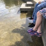 Zwei Personen sitzen auf einem Steinsims und halten ihre Beine ins Wasser eines flachen Teichs. Die eine trägt Jeans und Turnschuhe, die andere ein geblümtes Kleid und Sandalen. Der Teich ist klar und hat einen kiesigen Grund. Im Hintergrund sind Gras und Menschen zu sehen.