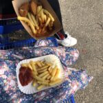 Zwei Personen sitzen auf einer blauen Metallbank. Die eine hält ein Tablett mit Pommes frites, Ketchup und Nuggets, die andere einen Pappteller mit Pommes frites, Ketchup und Mayonnaise. Die eine trägt einen geblümten Rock, die andere eine schwarze Hose und weiße Turnschuhe.