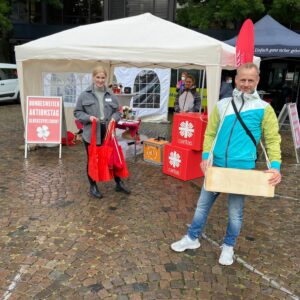 Zwei Personen stehen bei einer Veranstaltung im Freien vor einem weißen Zelt. Die eine hält rote Einkaufstaschen, die andere eine Holzkiste in der Hand. Um das Zelt herum sind rote Schilder und Informationsdisplays zu sehen.