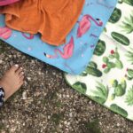 Ein nackter Fuß mit lackierten Zehennägeln steht auf dem Kies neben zwei bunten Handtüchern - einem blauen mit rosa Flamingos, einem weißen mit grünen Kakteen - und einem orangefarbenen Handtuch, das das Flamingo-Handtuch teilweise verdeckt.