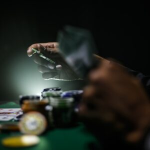 Nahaufnahme von Händen an einem Pokertisch, eine hält Spielkarten, die andere bereitet sich darauf vor, einen grünen Pokerchip zu platzieren. Pokerchips und Karten sind unter dramatischer Beleuchtung auf der Filzoberfläche verstreut.