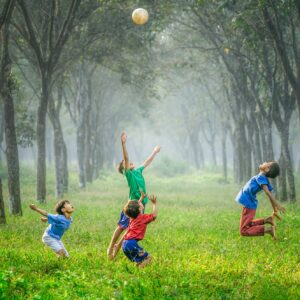 Vier Kinder spielen und springen, um einen Ball in einem grasbewachsenen, von Bäumen gesäumten Außenbereich zu erreichen, wobei das Sonnenlicht durch die Bäume und den nebligen Hintergrund fällt.