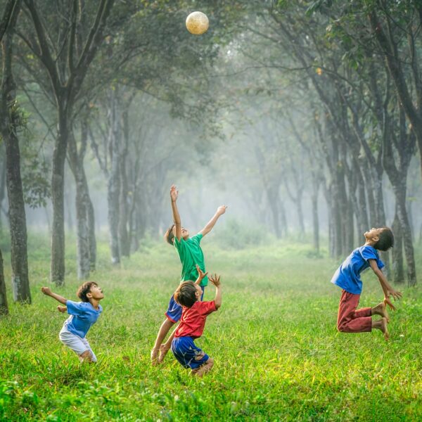 Vier Kinder spielen und springen, um einen Ball in einem grasbewachsenen, von Bäumen gesäumten Außenbereich zu erreichen, wobei das Sonnenlicht durch die Bäume und den nebligen Hintergrund fällt.