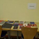 Ein Holztisch mit drei Stühlen enthält verschiedene Bücher und Materialien, die in zwei Abteilungen mit der Bezeichnung Methodenbeispiele und Literaturtisch untergebracht sind. Der Tisch steht vor einer beigen Wand.