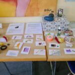 Auf einem Holztisch liegen verschiedene Gegenstände und bunte Karten mit Zeichnungen. Zu den Gegenständen gehören Stressbälle, ein Rubik-Würfel, Zappeltiere, Klebeband, kleine Behälter und bedruckte Blätter, die alle fein säuberlich angeordnet sind. Zwei Stühle stehen in der Nähe.