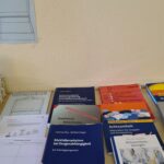 Ein Tisch mit verschiedenen Psychologie- und Therapiehandbüchern, Arbeitsblättern und Büchern, darunter Titel über dialektische Verhaltenstherapie und Achtsamkeit, unter einem Wandschild mit der Aufschrift "Methodenbeispiele" (auf Deutsch).