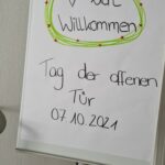Ein Flipchart mit einem handgeschriebenen Schild mit der Aufschrift "Herzlich Willkommen Tag der offenen Tür 07.10.2021" in deutscher Sprache, verziert mit einem grünen und roten Rand und einem gezeichneten Herz.