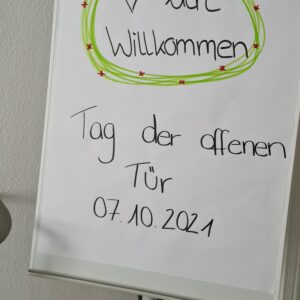 Ein Flipchart mit einem handgeschriebenen Schild mit der Aufschrift "Herzlich Willkommen Tag der offenen Tür 07.10.2021" in deutscher Sprache, verziert mit einem grünen und roten Rand und einem gezeichneten Herz.