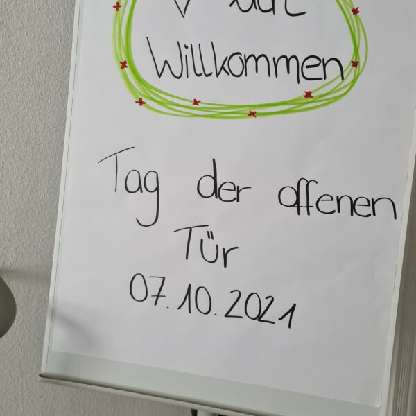 Ein Flipchart mit einem handgeschriebenen Schild mit der Aufschrift "Herzlich Willkommen Tag der offenen Tür 07.10.2021" in deutscher Sprache, verziert mit einem grünen und roten Rand und einem gezeichneten Herz.