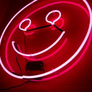 Ein neonpinkfarbenes Smiley-Schild leuchtet vor einer dunklen Wand, von einem niedrigen Winkel aus betrachtet. Der runde Umriss, die Augen und das breite Lächeln des Smileys sind hell erleuchtet.