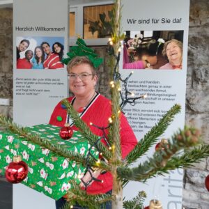 Eine lächelnde Frau mit einem festlichen Stirnband hält ein grünes Weihnachtsgeschenk neben einem geschmückten Baum mit Lichtern. Hinter ihr sind Begrüßungsplakate in deutscher Sprache und Fotos von Menschen zu sehen.