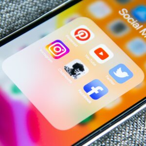 Auf dem Bildschirm eines Smartphones wird ein Ordner mit der Aufschrift Social Media mit Symbolen für Pinterest, Instagram, Clubhouse, YouTube, Twitter und Facebook vor einem bunten, verschwommenen Hintergrund angezeigt.