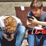 Zwei Teenager sitzen im Freien auf einem Ziegelstein, beide schauen auf ihre Smartphones. Im Hintergrund sitzen Menschen auf Bänken. Die Worte klar sein erscheinen in weißer Schrift auf einem roten Pinselstrich.