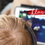 Ein Kind mit blondem Haar und Kopfhörern benutzt ein Tablet. Die Worte klar sein erscheinen in Kursivschrift auf einem roten Pinselstrich in der oberen rechten Ecke.