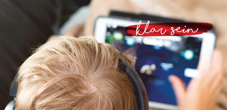Ein Kind mit blondem Haar und Kopfhörern benutzt ein Tablet. Die Worte klar sein erscheinen in Kursivschrift auf einem roten Pinselstrich in der oberen rechten Ecke.