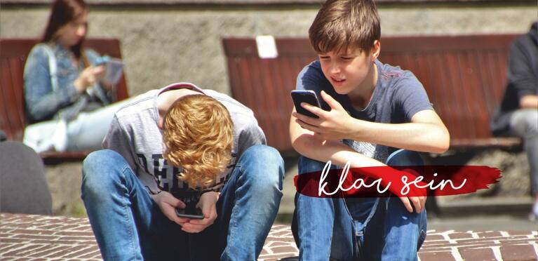 Zwei Teenager sitzen im Freien auf einem Ziegelstein, beide schauen auf ihre Smartphones. Im Hintergrund sitzen Menschen auf Bänken. Die Worte klar sein erscheinen in weißer Schrift auf einem roten Pinselstrich.
