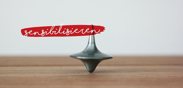 Ein Kreisel balanciert auf einer Holzfläche. Darüber steht in weißer Handschrift auf einem roten Farbstreifen das deutsche Wort sensibilisieren. Der Hintergrund ist einfarbig und hell gefärbt.