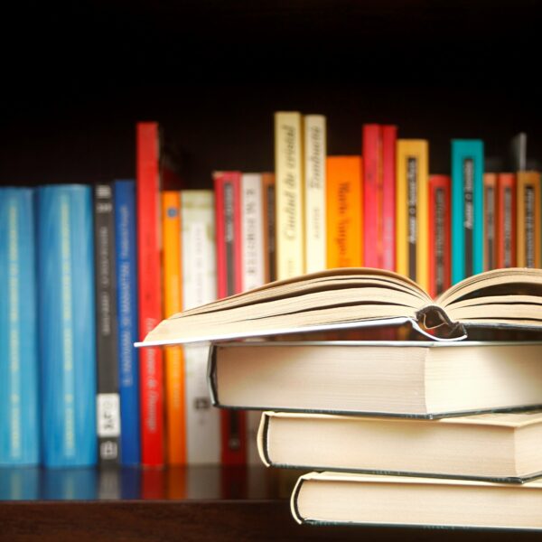 Ein Stapel von drei Büchern liegt auf einem Tisch, ein aufgeschlagenes Buch liegt obenauf. Dahinter sind bunte Bücher fein säuberlich in einem Bücherregal angeordnet.
