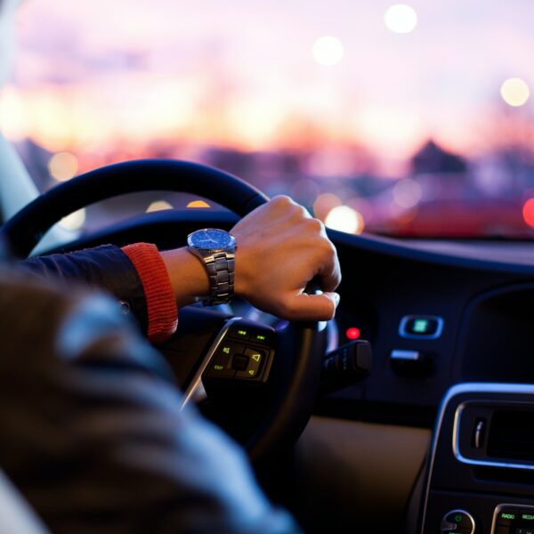 Eine Person, die eine Uhr und eine Lederjacke trägt, fährt bei Sonnenuntergang in einem Auto, wobei das Armaturenbrett und das Lenkrad sichtbar sind und die Lichter der Stadt außerhalb des Fensters verschwimmen.