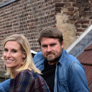 Eine lächelnde Frau mit blondem Haar und ein Mann mit braunem Haar und Bart sitzen im Freien vor einem Backsteingebäude und einem Ziegeldach, beide in Freizeitkleidung.