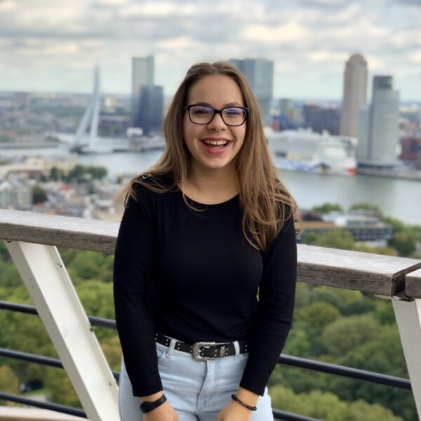 Eine junge Frau mit langen braunen Haaren, Brille, schwarzem Oberteil und hellblauen Jeans lächelt, während sie auf einem Balkon steht und die Skyline einer Stadt mit hohen Gebäuden, einem Fluss und einer Brücke im Hintergrund überblickt.