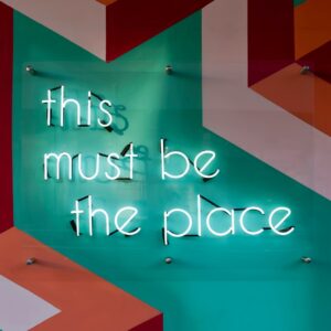 Ein Neonschild mit der Aufschrift "This must be the place" ist an einer farbenfrohen, geometrisch gemusterten Wand mit türkisfarbenen, orangefarbenen, rosa und weißen Formen angebracht.