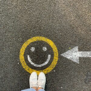 Person mit weißen Schuhen und hellblauer Hose, die vor einem gelben, auf Asphalt gemalten Smiley steht, neben dem ein weißer Pfeil nach rechts zeigt.