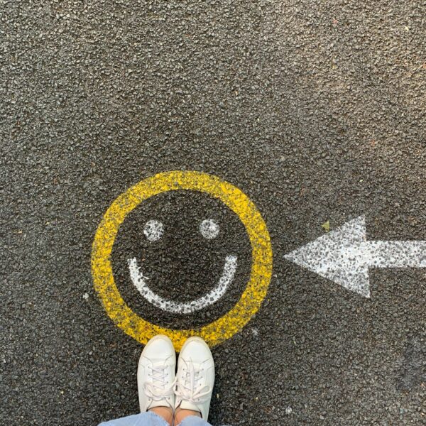 Person mit weißen Schuhen und hellblauer Hose, die vor einem gelben, auf Asphalt gemalten Smiley steht, neben dem ein weißer Pfeil nach rechts zeigt.