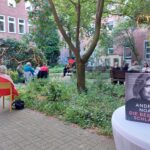 Eine Gruppe von Menschen sitzt auf Stühlen in einem Gartenhof, wo eine Veranstaltung im Freien stattfindet. Im Vordergrund liegt ein Buch mit dem Titel Die Bestie Schläft von Andrea Noack auf einem runden weißen Tisch. Lichterketten hängen in den Bäumen.