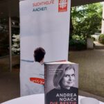 Ein Buch mit dem Titel DIE BESTIE SCHLÄFT von Andrea Noack steht auf einem weißen Tisch neben einer weißen Tasse. Dahinter ist ein Banner der Suchthilfe Aachen und der Caritas zu sehen, mit einer Außenanlage und Grünpflanzen.