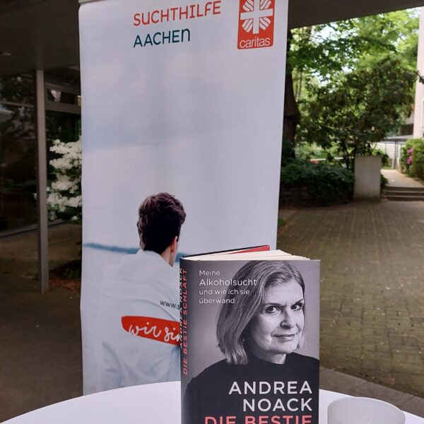 Ein Buch mit dem Titel "Die Bestie schläft" von Andrea Noack steht auf einem Tisch im Freien, vor einem Banner der Caritas Suchthilfe Aachen. Im Hintergrund sind Bäume und ein Gebäude zu sehen.